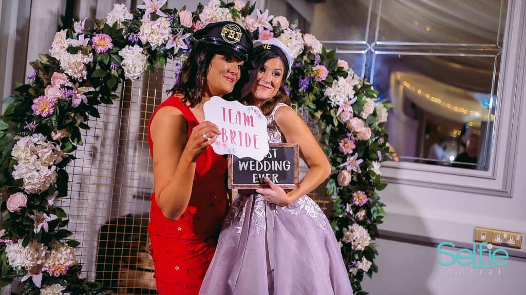 Two women at a wedding photo booth, one holding a "Team Bride" sign, smiling in front of a floral ar.