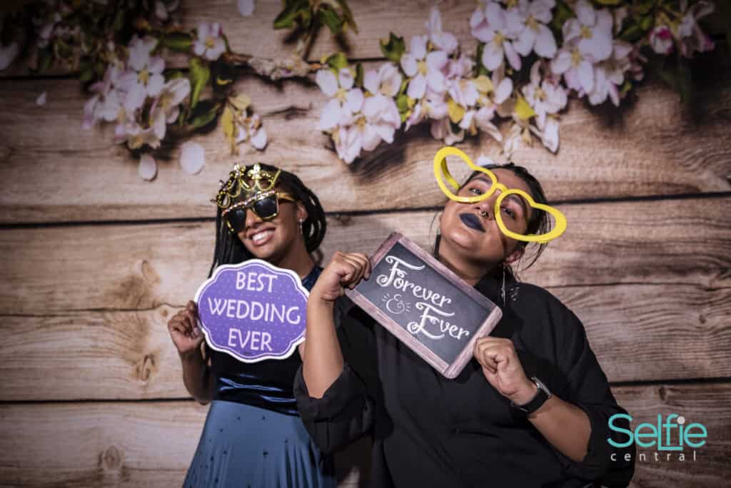 Two women enjoying a photo booth at an event, wearing fun accessories and holding signs.