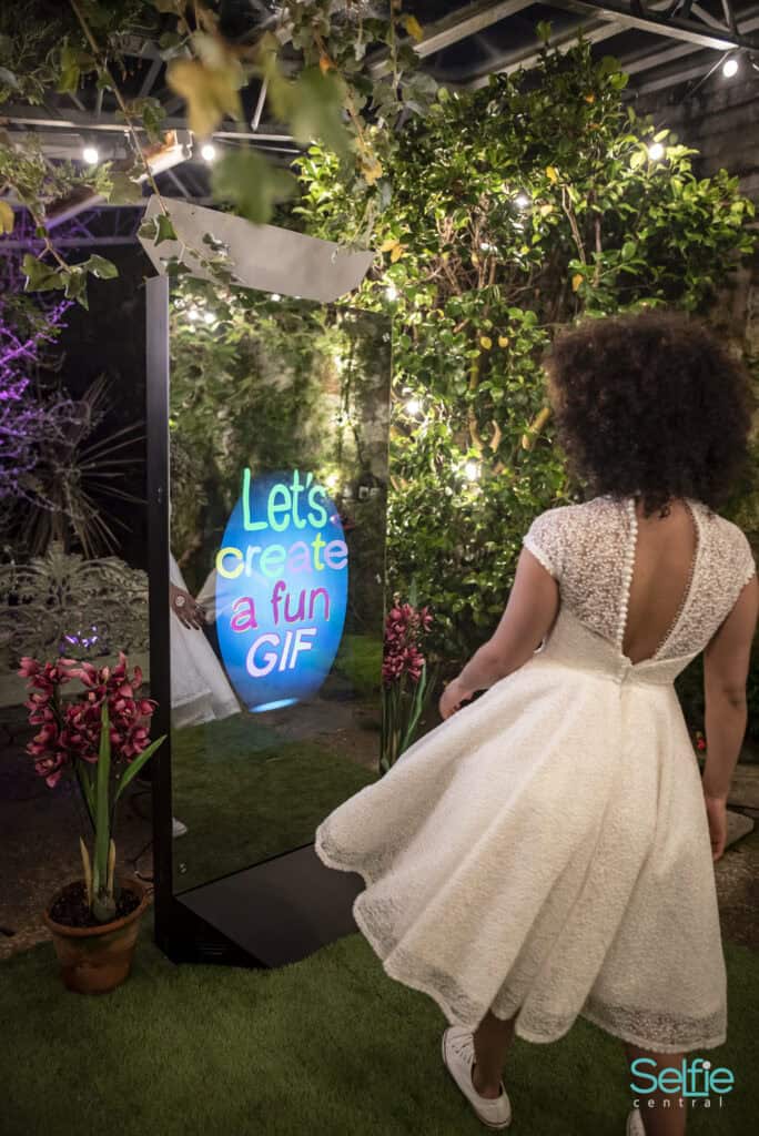 Woman using a selfie mirror at an event with vibrant greenery and neon sign.