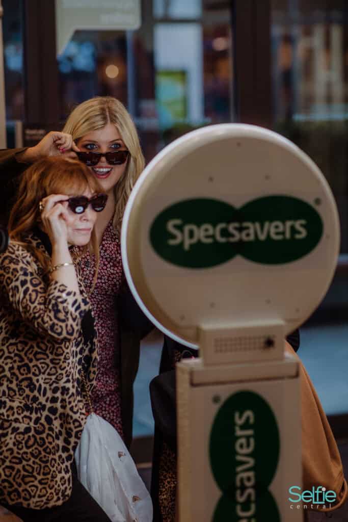 Two women using a photo booth at an event in Auckland, smiling and wearing sunglasses.